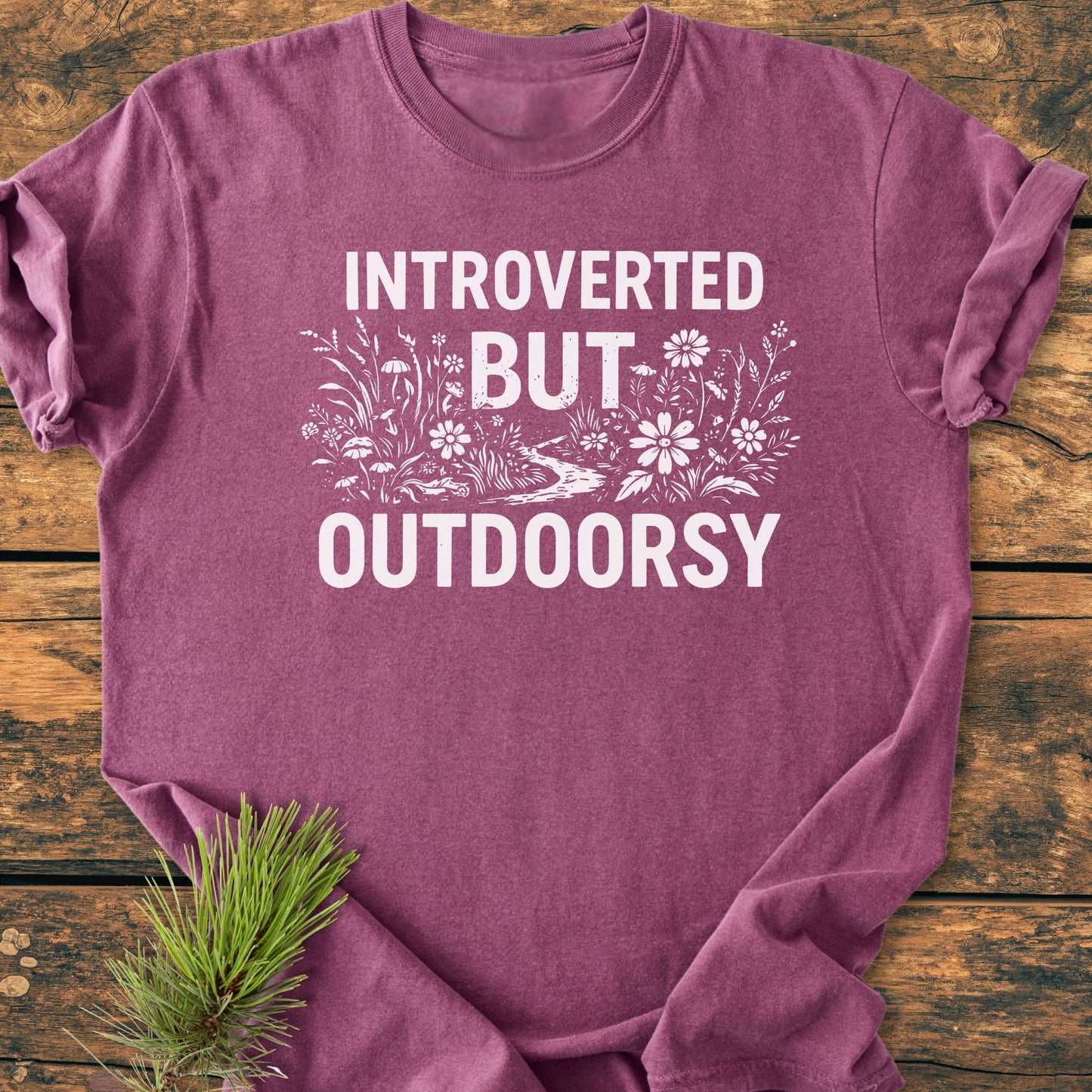 Introverted But Outdoorsy - Vintage Wash Tee features bold white text and wildflowers on a heathered plum-purple shirt, laid on wood with a pine sprig accentuating its nature-inspired theme.