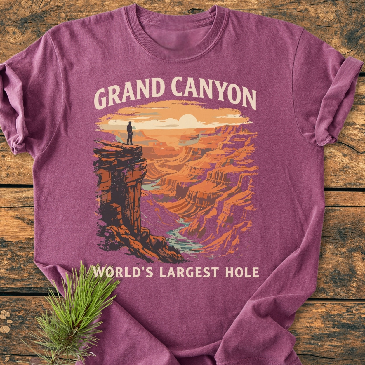Grand Canyon Large Hole - Vintage Wash Tee on wood, featuring a retro Grand Canyon print at sunrise/sunset with a lone figure. Adjacent is a pine sprig, emphasizing its nature-inspired theme.