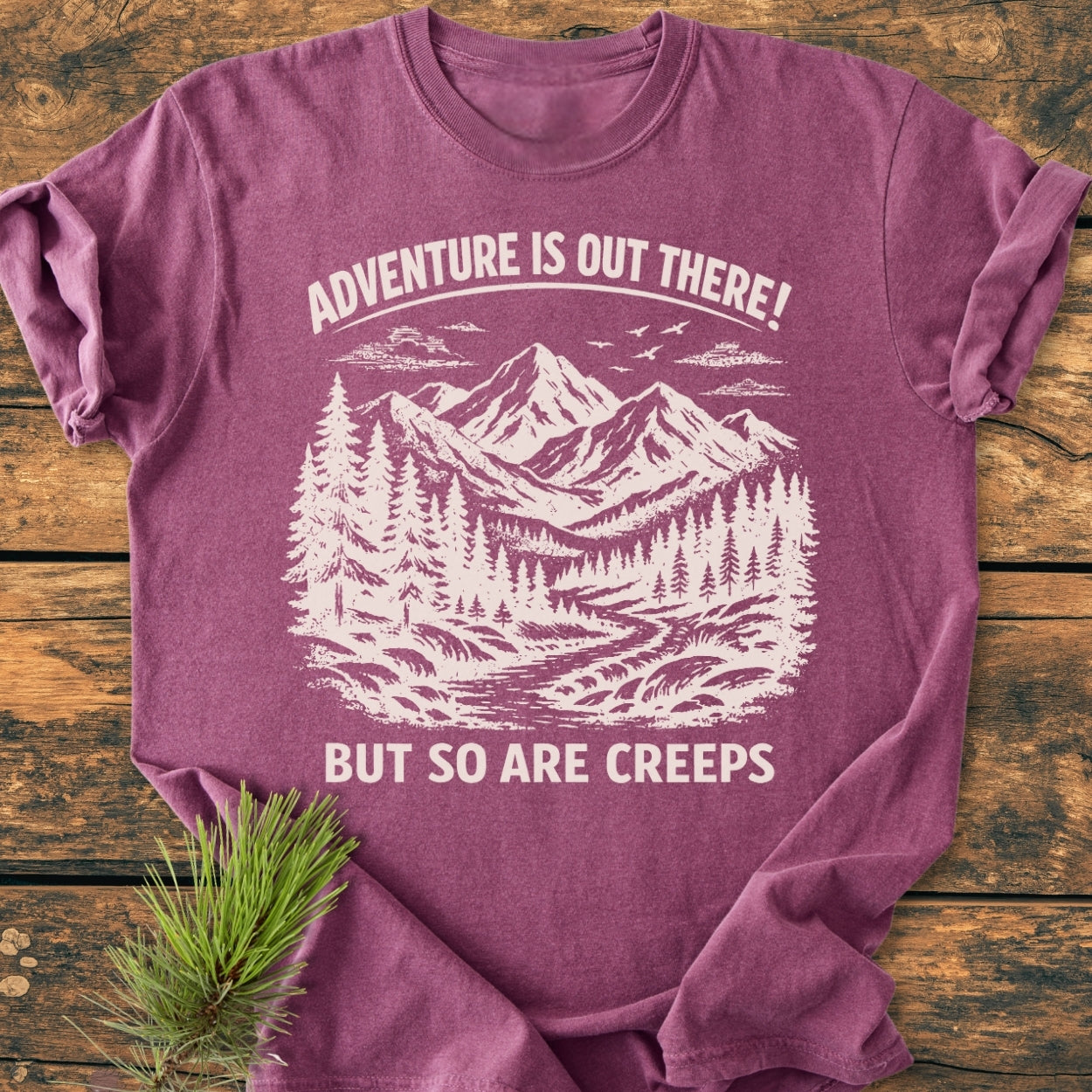 Adventure Is Out There - Vintage Wash Tee featuring a stylized wilderness scene in off-white ink on a heathered plum T-shirt, laid on wood with a pine sprig accentuating its rustic charm.