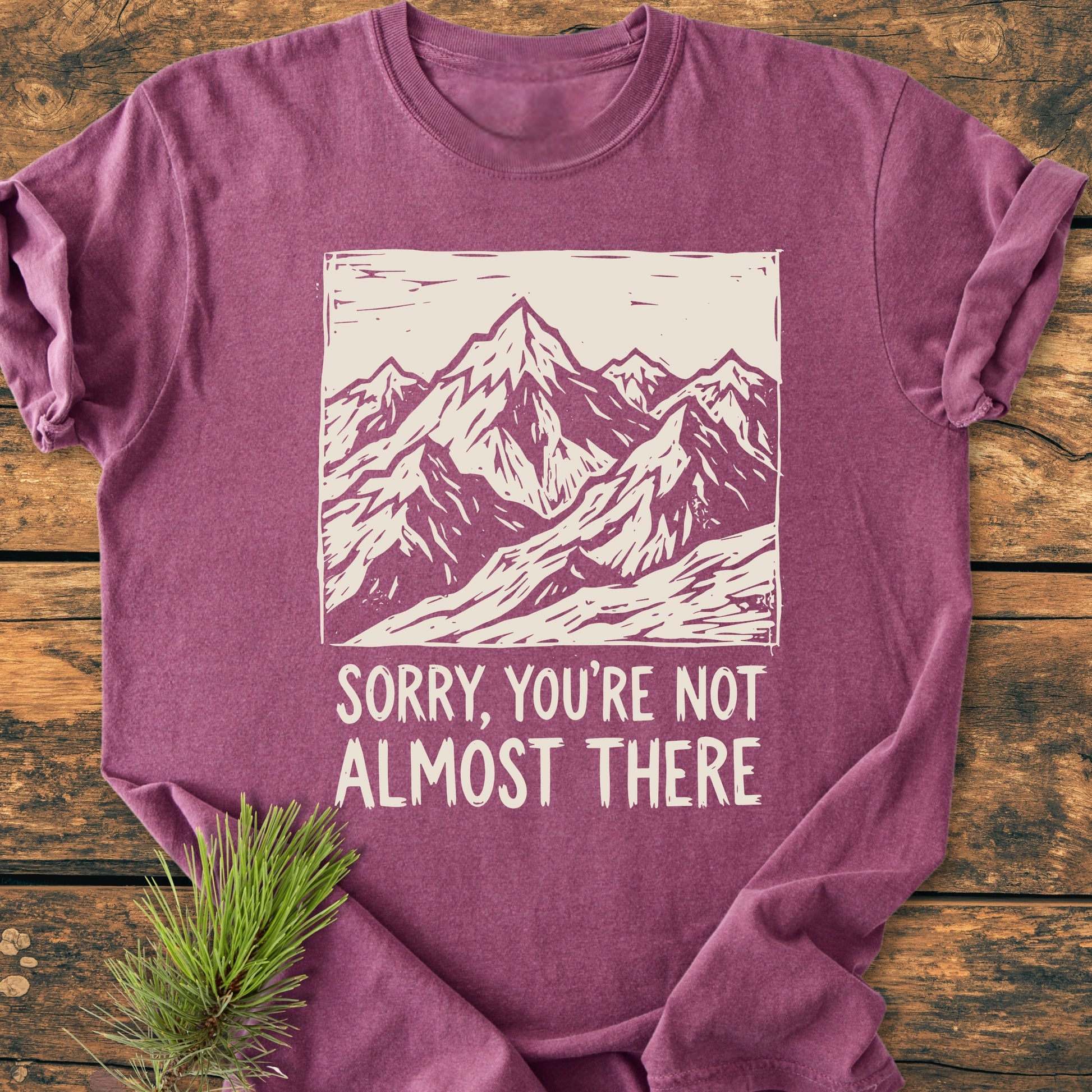 Not Almost There - Vintage Wash Tee features a plum hue with a cream mountain design and phrase. Laid on wood, an evergreen sprig accents this soft, durable, garment-dyed, ring-spun cotton shirt.