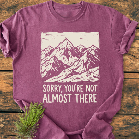 Not Almost There - Vintage Wash Tee features a plum hue with a cream mountain design and phrase. Laid on wood, an evergreen sprig accents this soft, durable, garment-dyed, ring-spun cotton shirt.