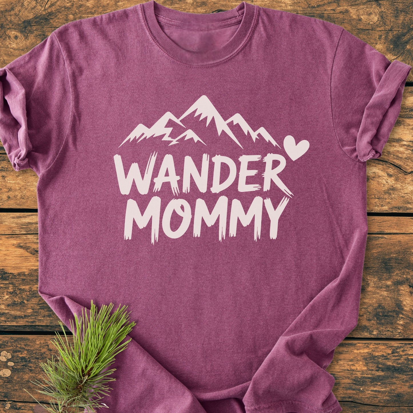 Wander Mommy - Vintage Heavyweight Tee featuring a white mountain and heart graphic on a plum-purple shirt, laid flat on wood with a pine sprig, showcasing its relaxed, durable design.