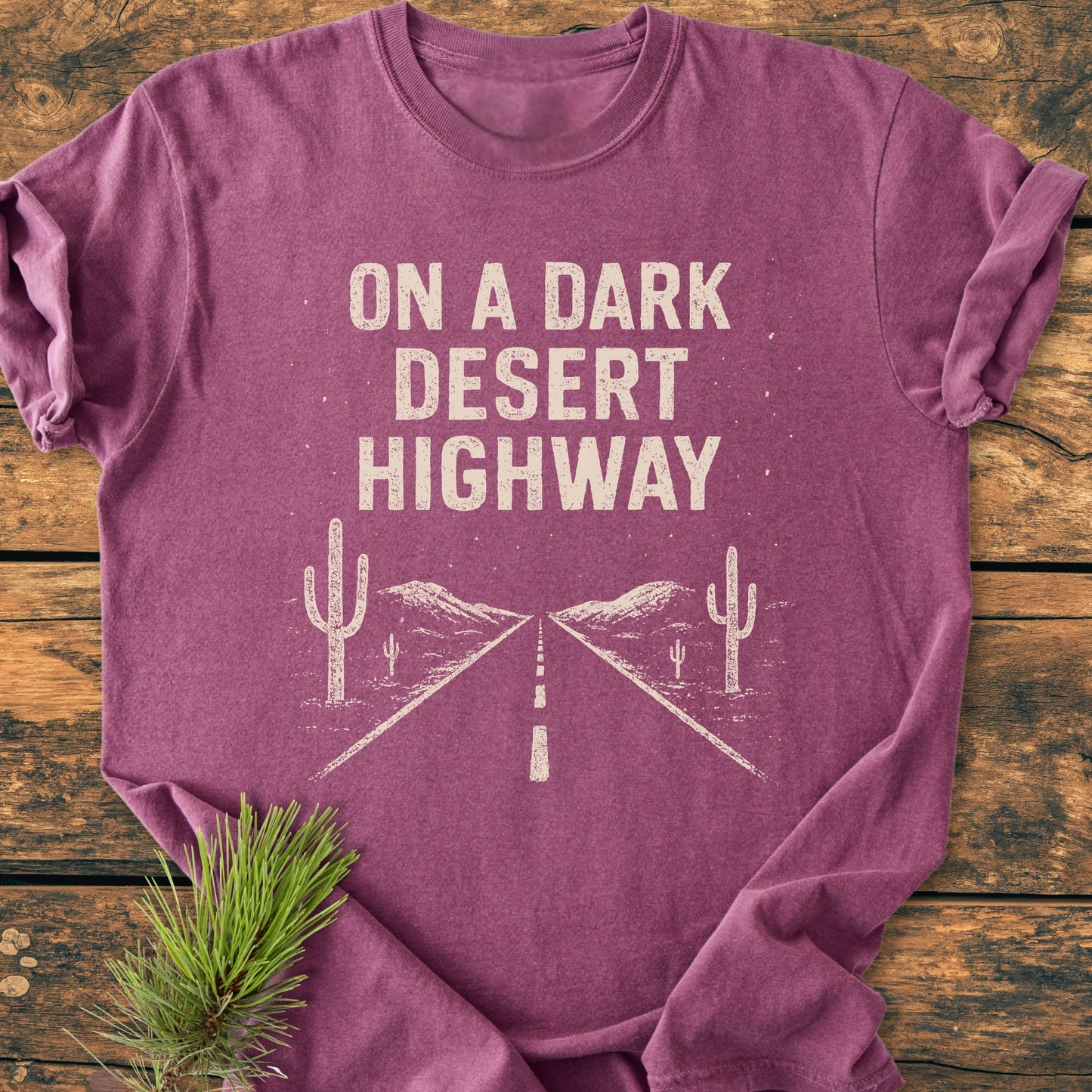 On A Dark Desert Highway - Vintage Wash Tee featuring off-white text and desert graphic, laid flat on rustic wood with pine sprig, embodying a relaxed unisex fit and vintage feel.
