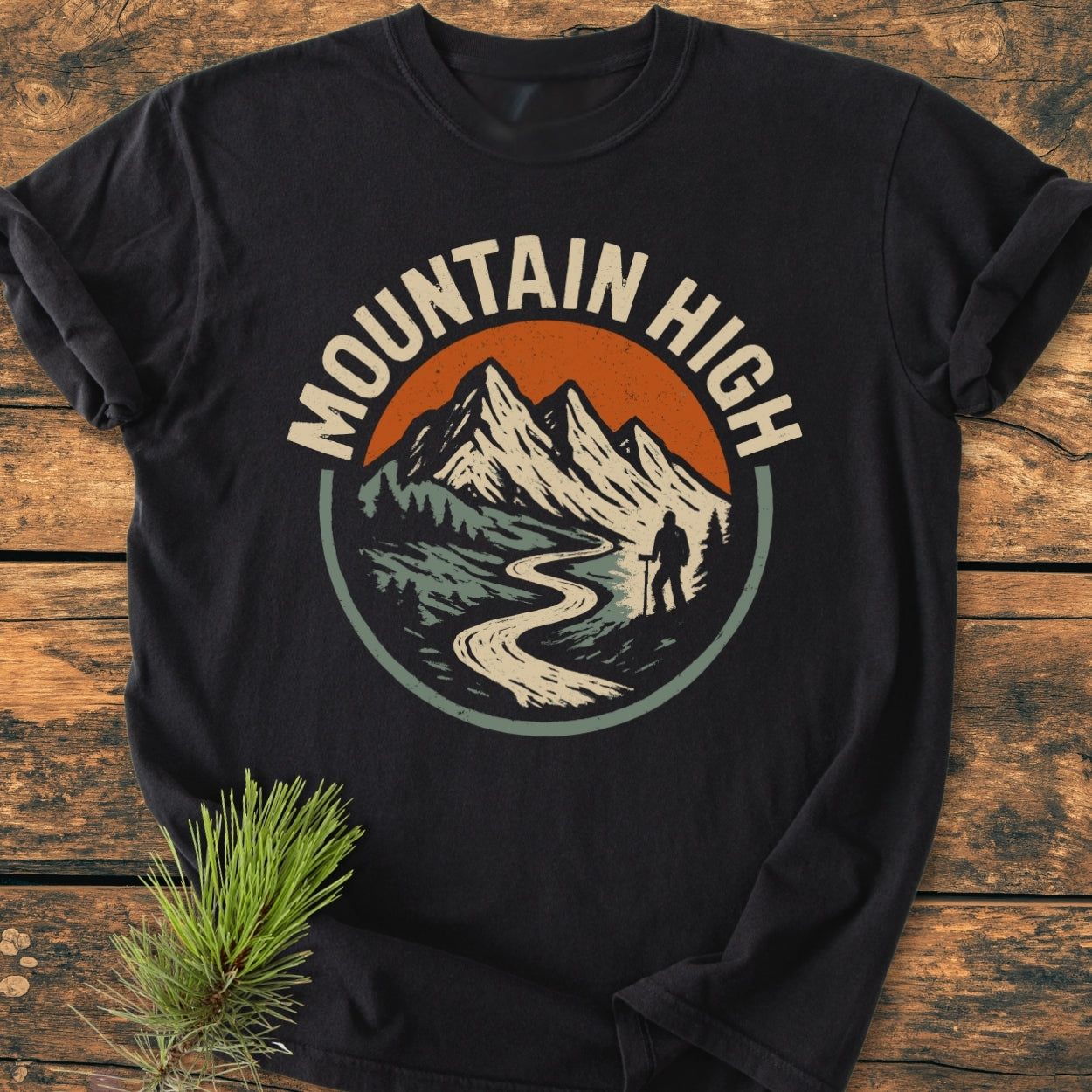 Mountain High - Vintage Wash Tee: Black T-shirt featuring a mountain graphic with a hiker and MOUNTAIN HIGH text, laid on rustic wood with pine needles, showcasing its soft, relaxed fit and durable stitching.
