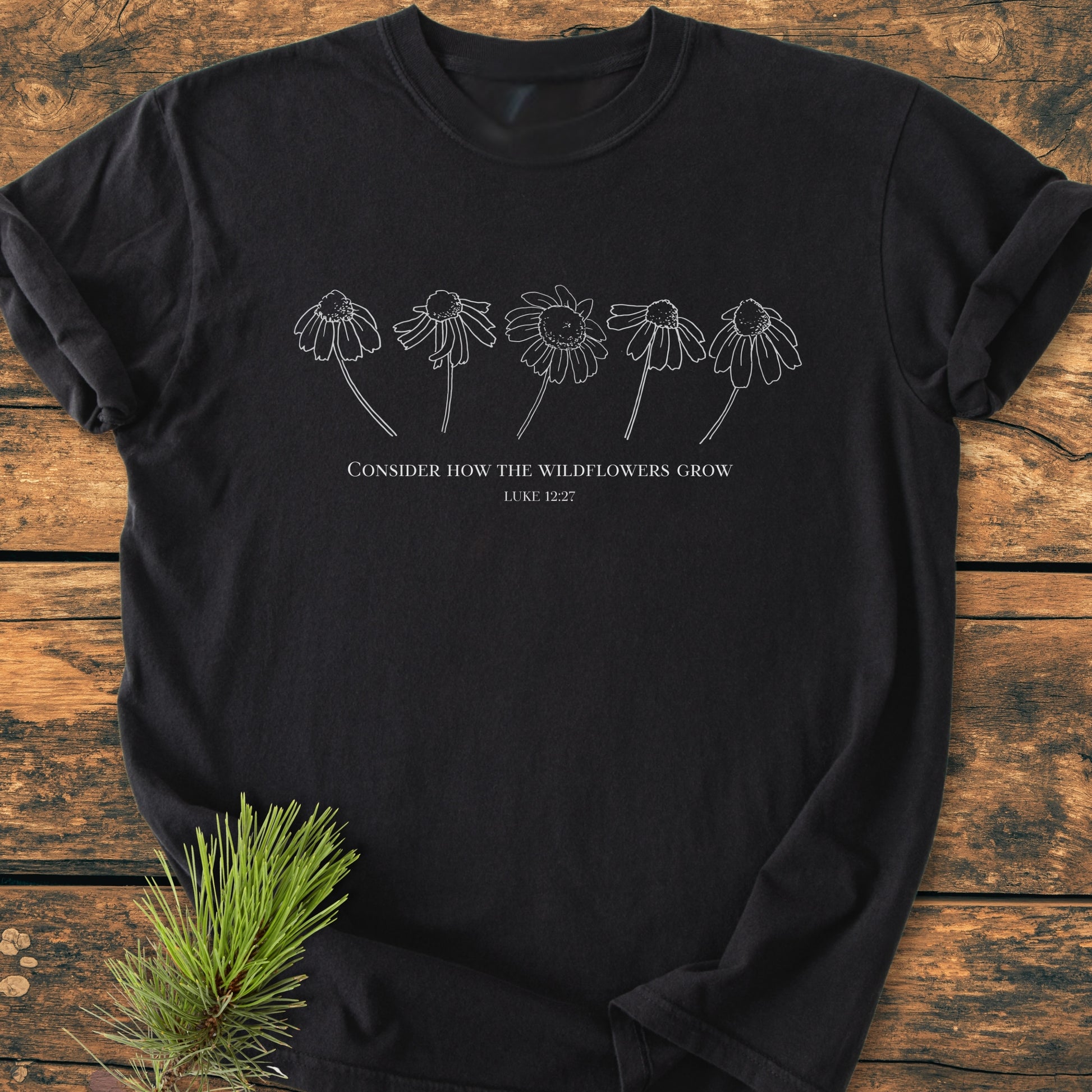 Consider How The Wildflowers Grow - Vintage Wash Tee features a white wildflower line-drawing with Luke 12:27 quote on a black t-shirt, placed on rustic wood, accented by a pine sprig.