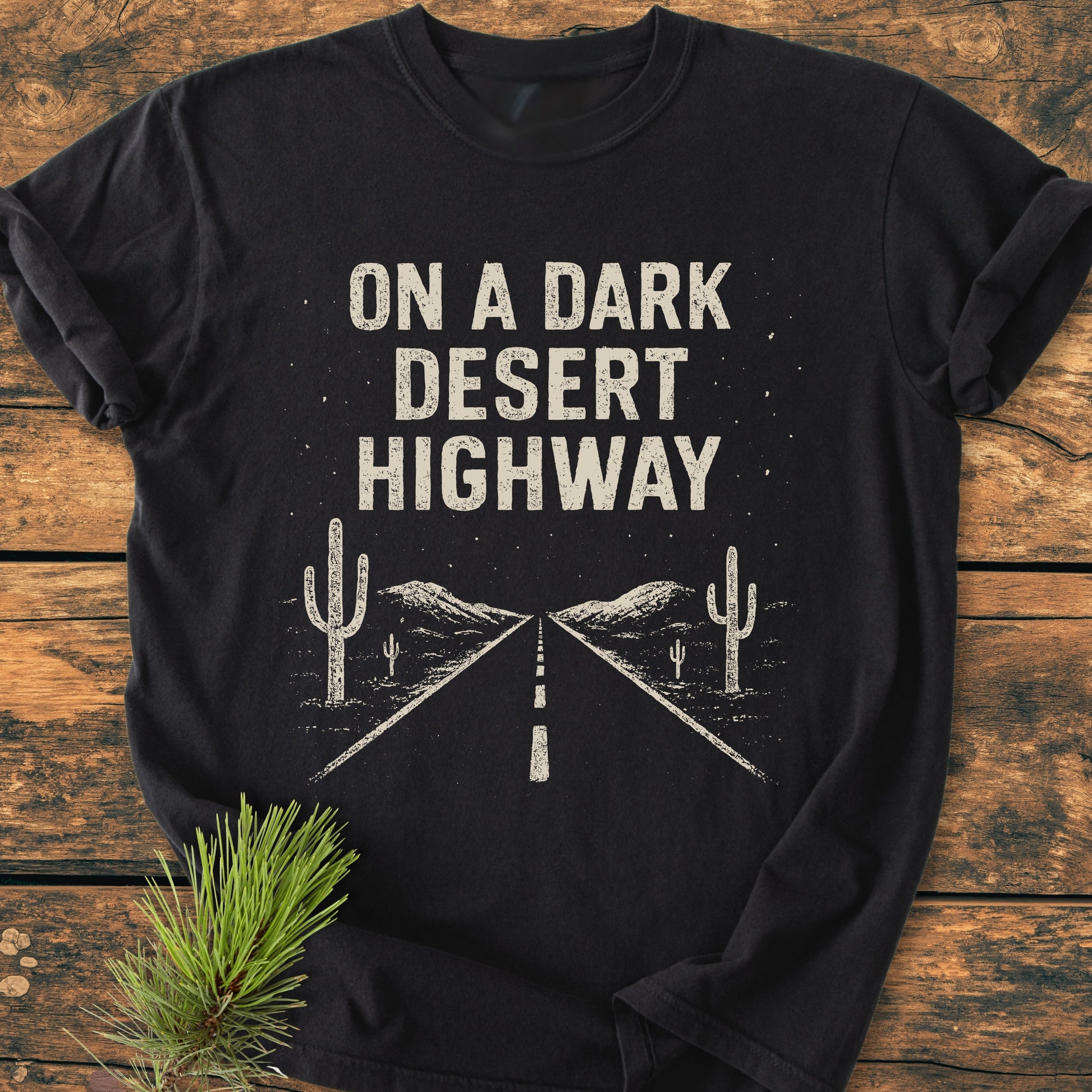 On A Dark Desert Highway - Vintage Wash Tee: Black long-sleeve tee with off-white desert scene print, laid on rustic wood, paired with a pine sprig, embodying a hiker's dream.