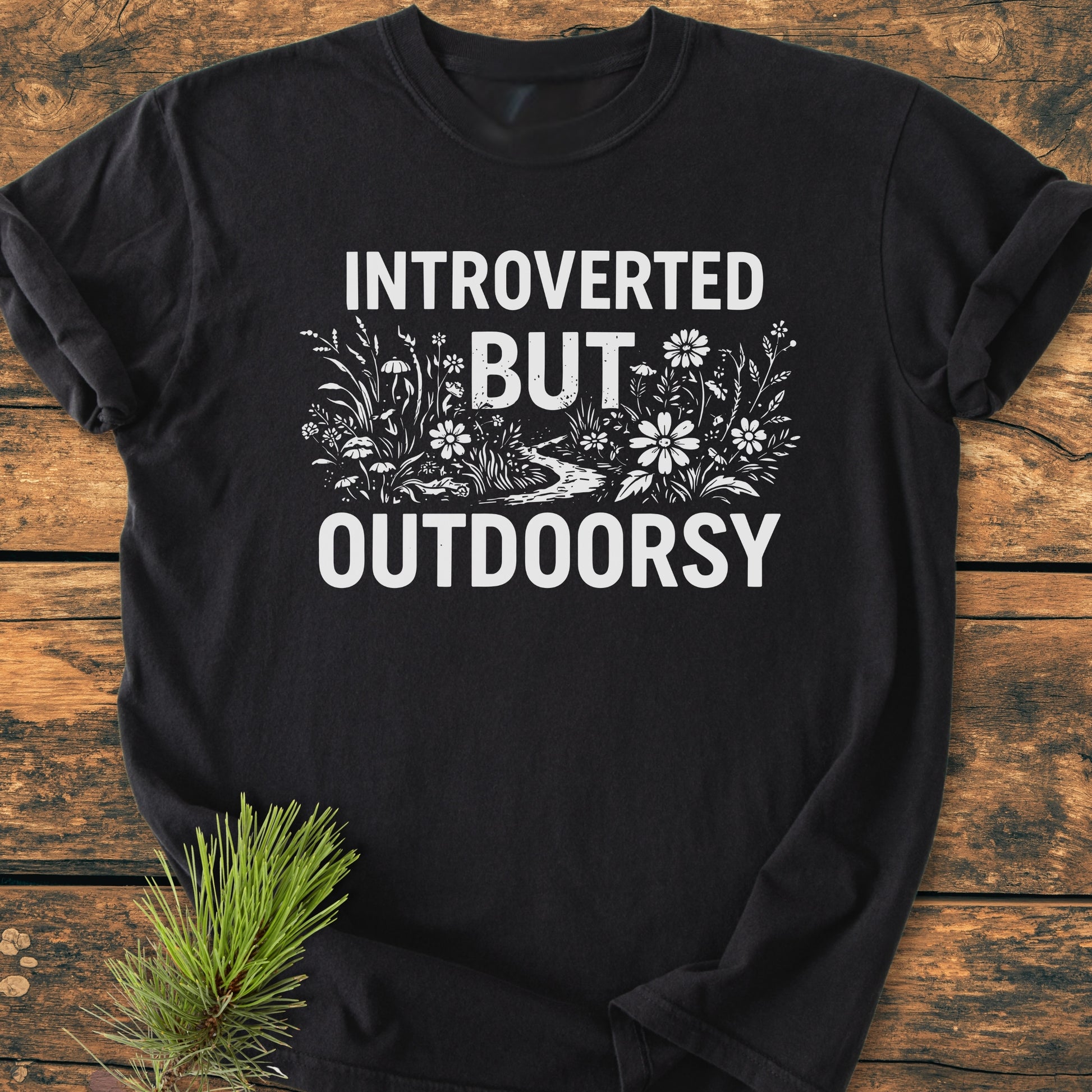 Introverted But Outdoorsy - Vintage Wash Tee displayed on a wooden surface, featuring bold white text and nature line-drawing. Includes soft, relaxed fit, emphasizing comfort with durable, seamless construction.