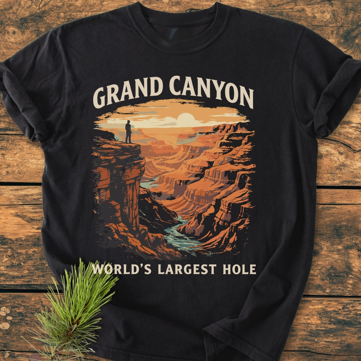 Grand Canyon Large Hole - Vintage Wash Tee features a sunset-lit Grand Canyon illustration on a black T-shirt, highlighting layered cliffs and a lone figure, laid on a wooden surface with a pine sprig.