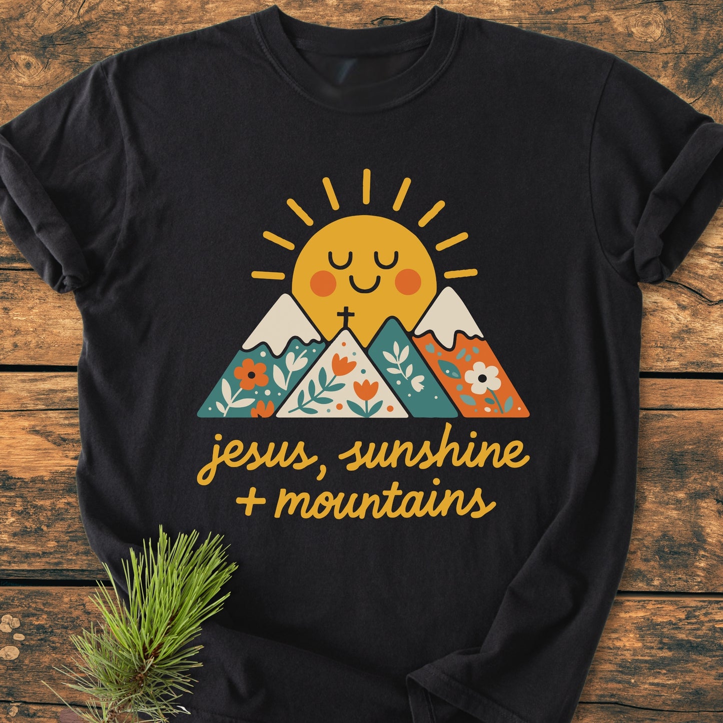 Jesus, Sunshine, Mountains - Vintage Heavyweight Tee featuring a smiling sun and floral-patterned mountains graphic. Hand-lettered phrase “jesus, sunshine + mountains” on front, laid on wooden surface.