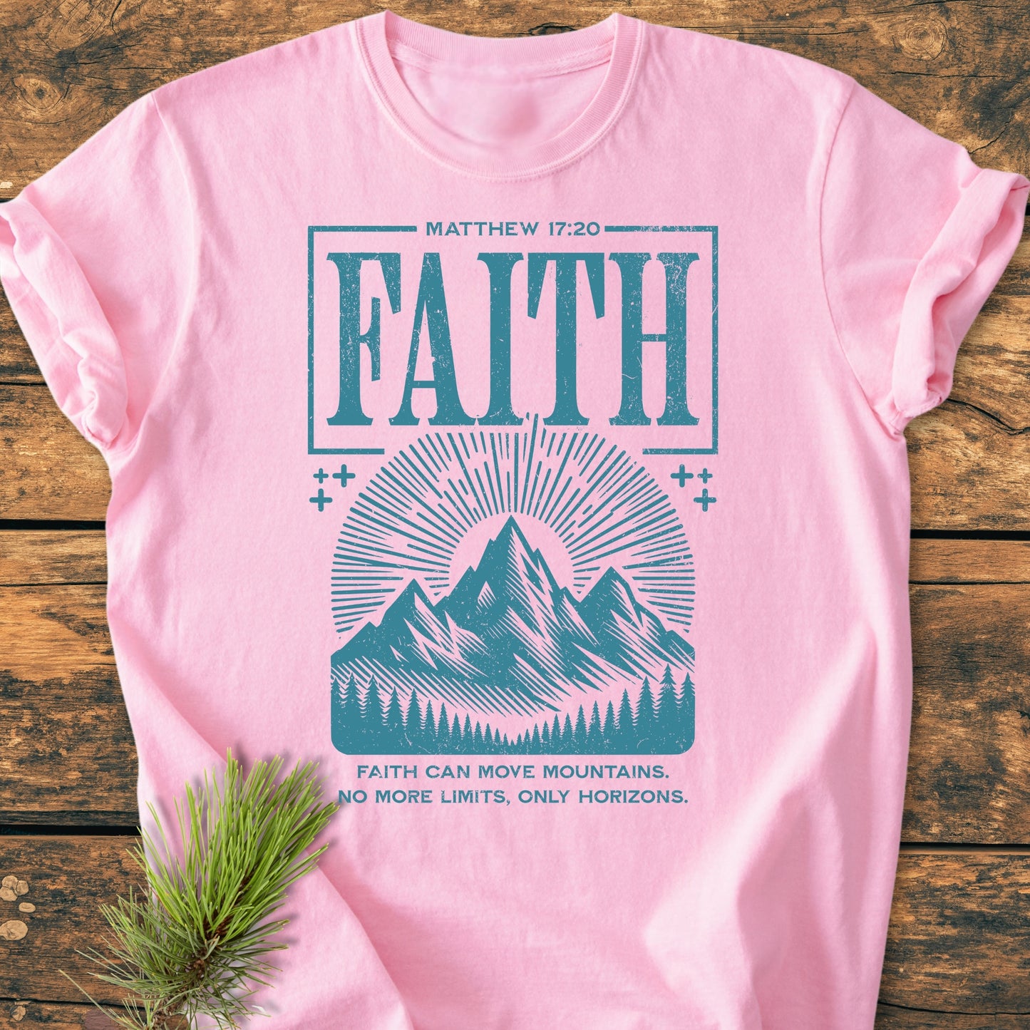 Faith Moves Mountains - Vintage Wash Tee: Soft pink T-shirt featuring a teal-blue MATTHEW 17:20 FAITH graphic with mountain illustration and slogan, laid on wood, pine sprig at corner.