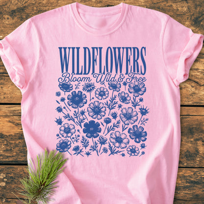 Wildflowers Bloom Wild And Free - Vintage Wash Tee featuring navy-blue WILDFLOWERS graphic with wildflower illustrations on a pink shirt, styled on a wooden surface with a pine sprig.