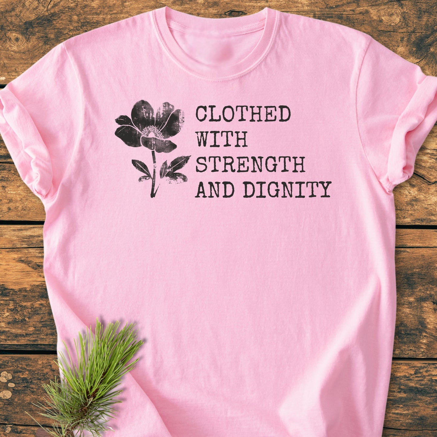 Soft pink Clothed With Strength And Dignity, Proverbs 31:25 - Vintage Wash Tee features a floral illustration and quote on the front, styled with a pine sprig on a wooden surface.