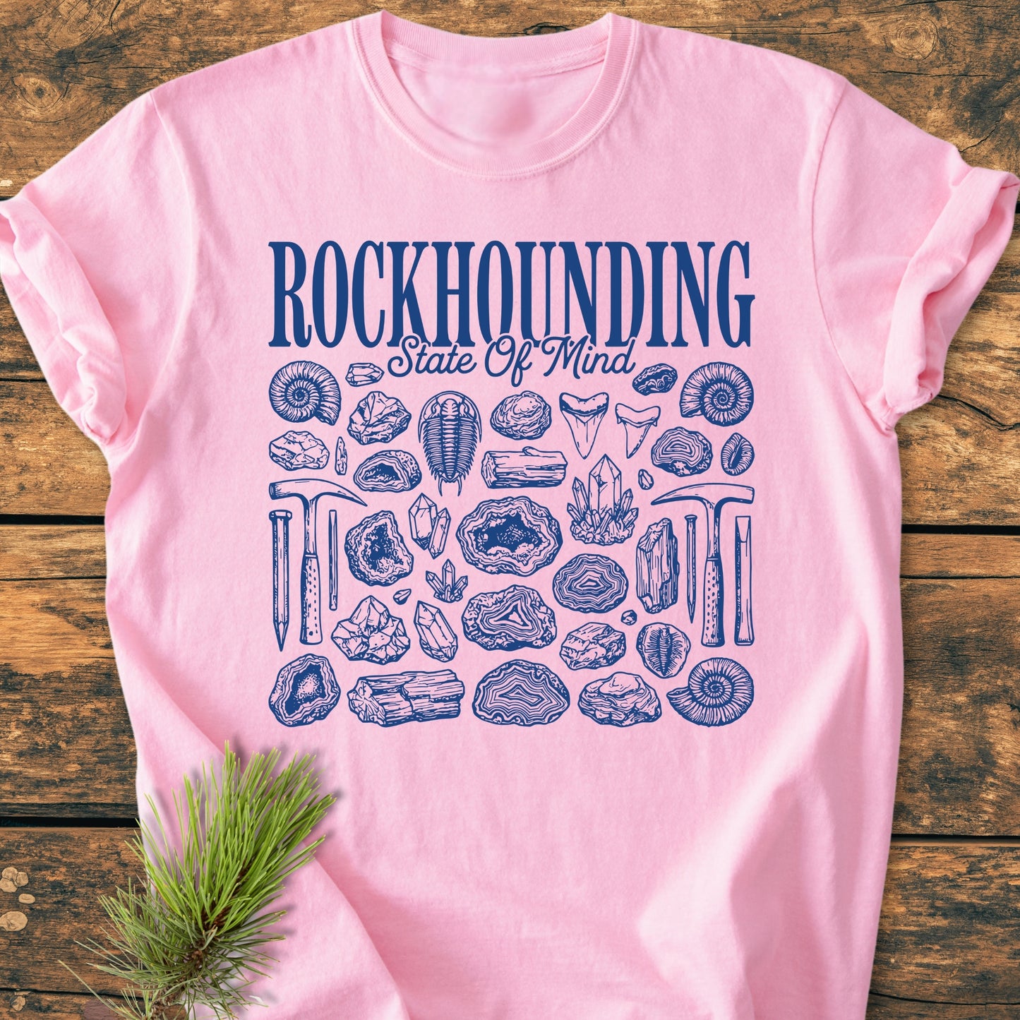 Rockhounding State Of Mind - Vintage Wash Tee: Soft-pink shirt with dark blue geological motifs, showcasing rocks, fossils, and tools. Laid on rustic wood, accented with a pine sprig, embodying earth science passion.
