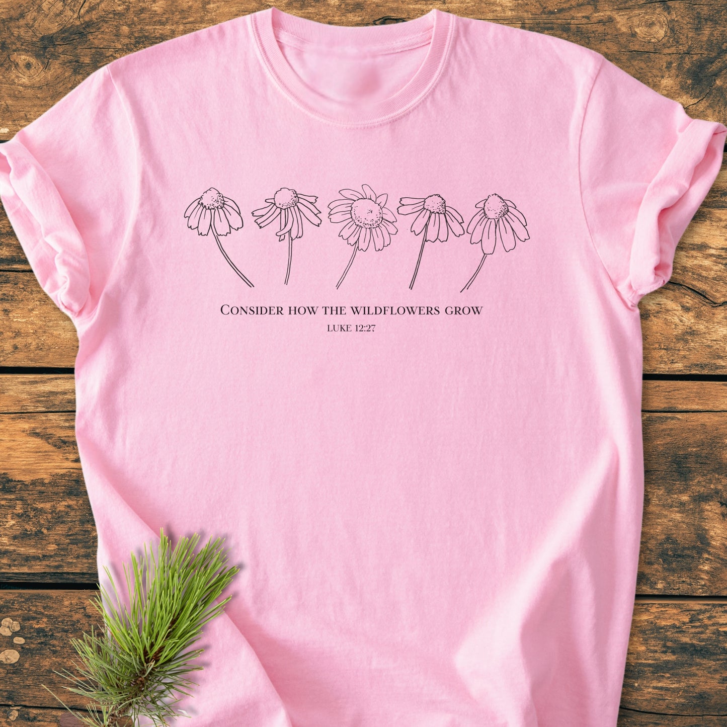 Consider How The Wildflowers Grow - Vintage Wash Tee features a soft-pink hue with wildflower illustrations and a biblical quote, laid flat on wood, emphasizing its cozy, relaxed fit and durable construction.