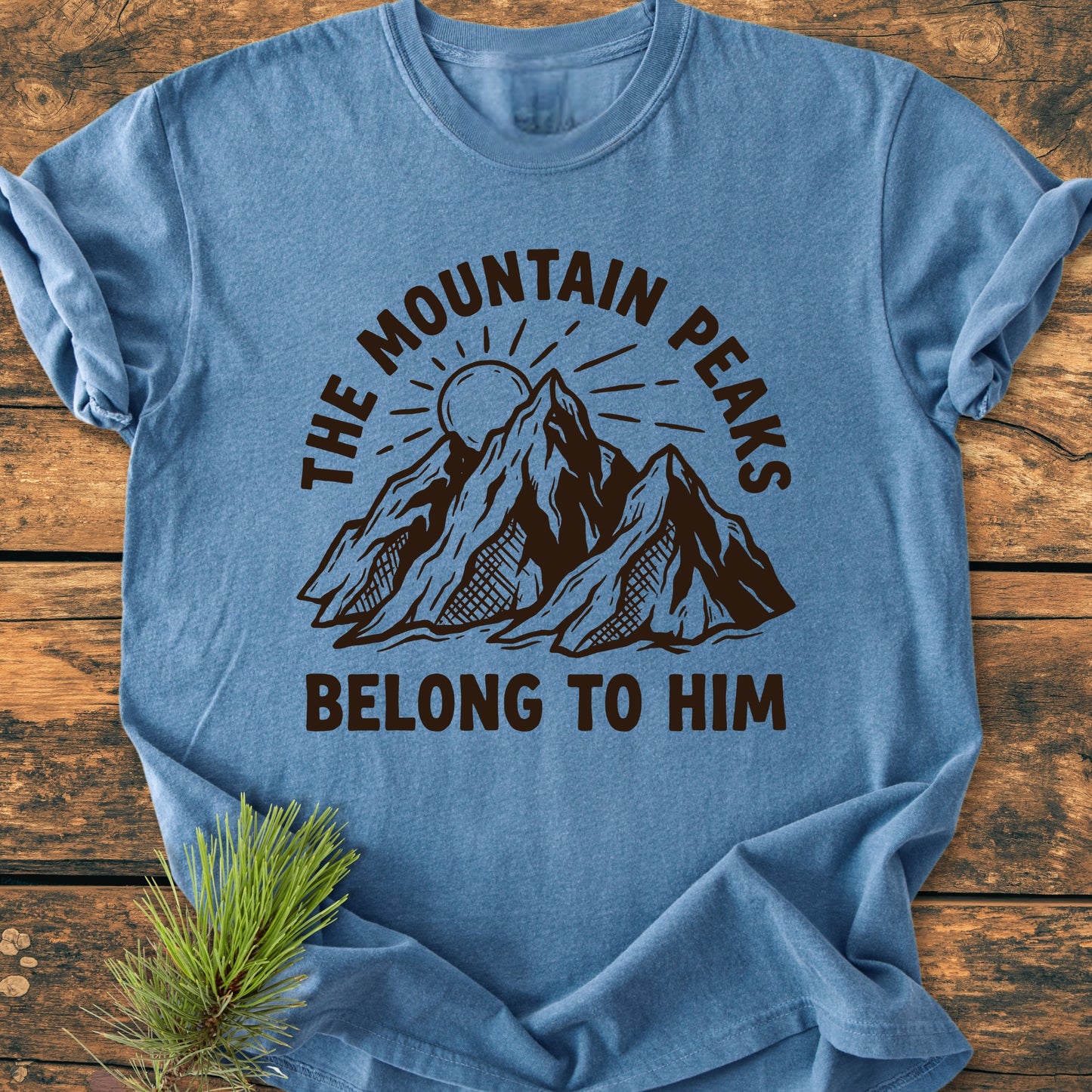 The Mountain Peaks Belong To Him - Vintage Wash Tee features a stylized mountain range and sun design, screen-printed on a crew-neck T-shirt, laid flat with a small pine sprig nearby.