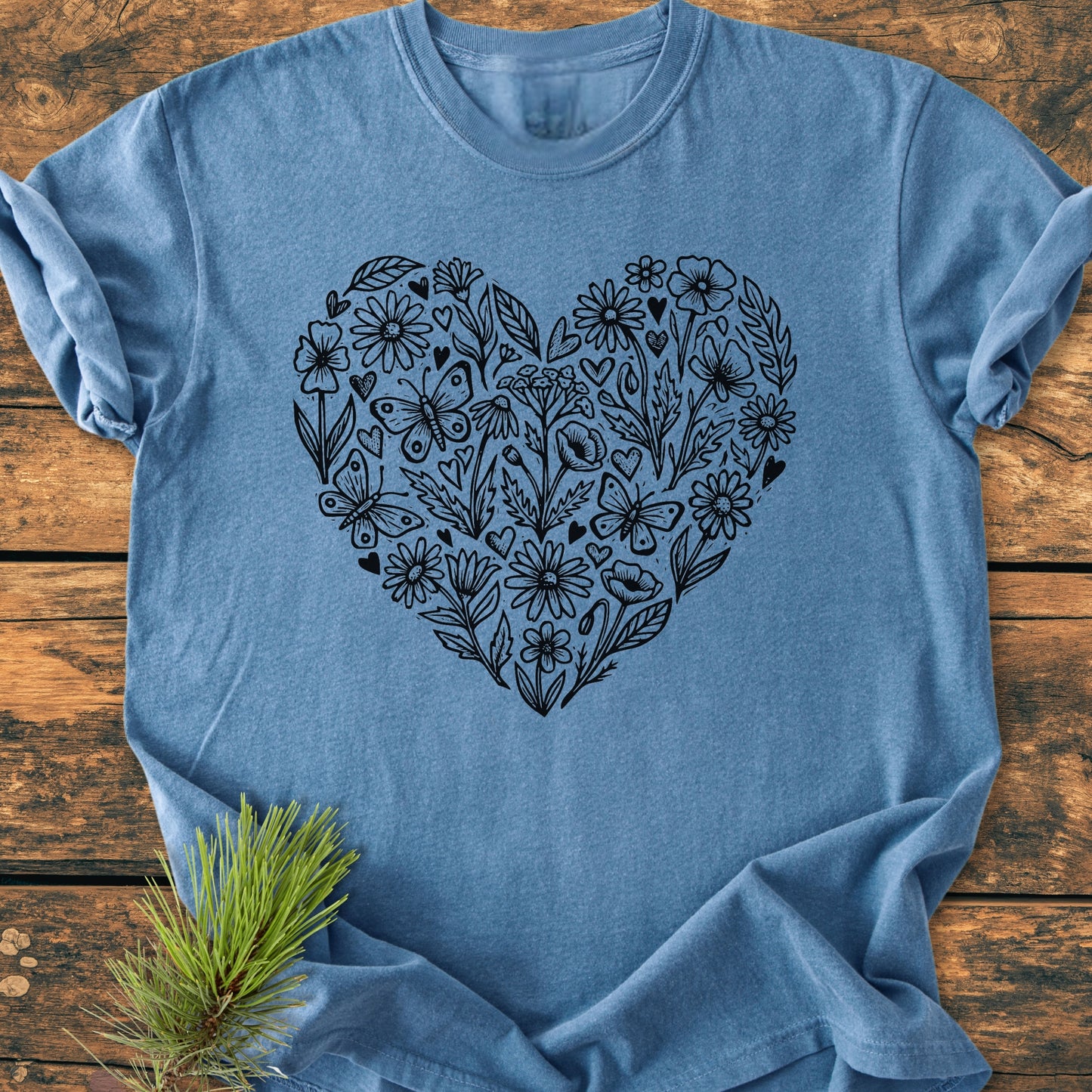 Heather-blue Wildflower Heart Butterfly Soul - Vintage Heavyweight Tee featuring a black line art heart with flowers, leaves, butterflies, and tiny hearts. Laid flat with casually rolled sleeves on a wooden surface.