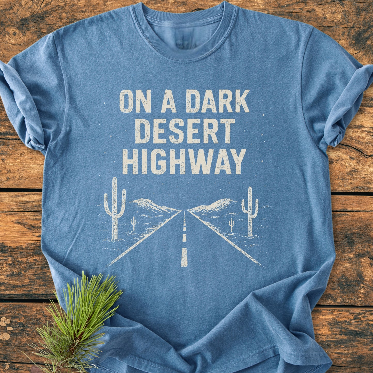 Flat-lay of the On A Dark Desert Highway - Vintage Wash Tee in heathered blue, featuring a desert road and cacti design, draped on wood with a pine branch accent.