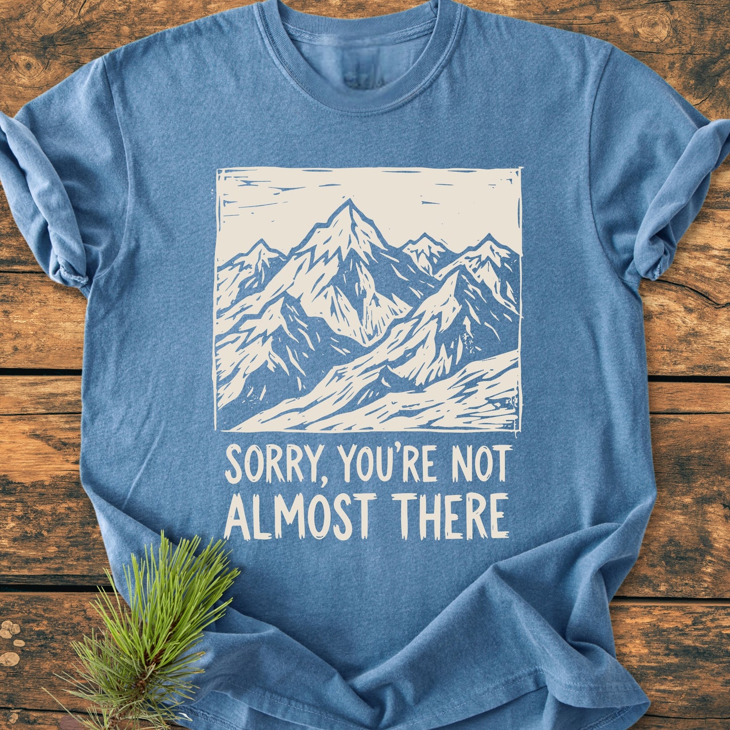 Not Almost There - Vintage Wash Tee featuring a hand-drawn mountain graphic and slogan. Light-blue, crew-neck design on a wooden surface, with a pine sprig accentuating its natural aesthetic.
