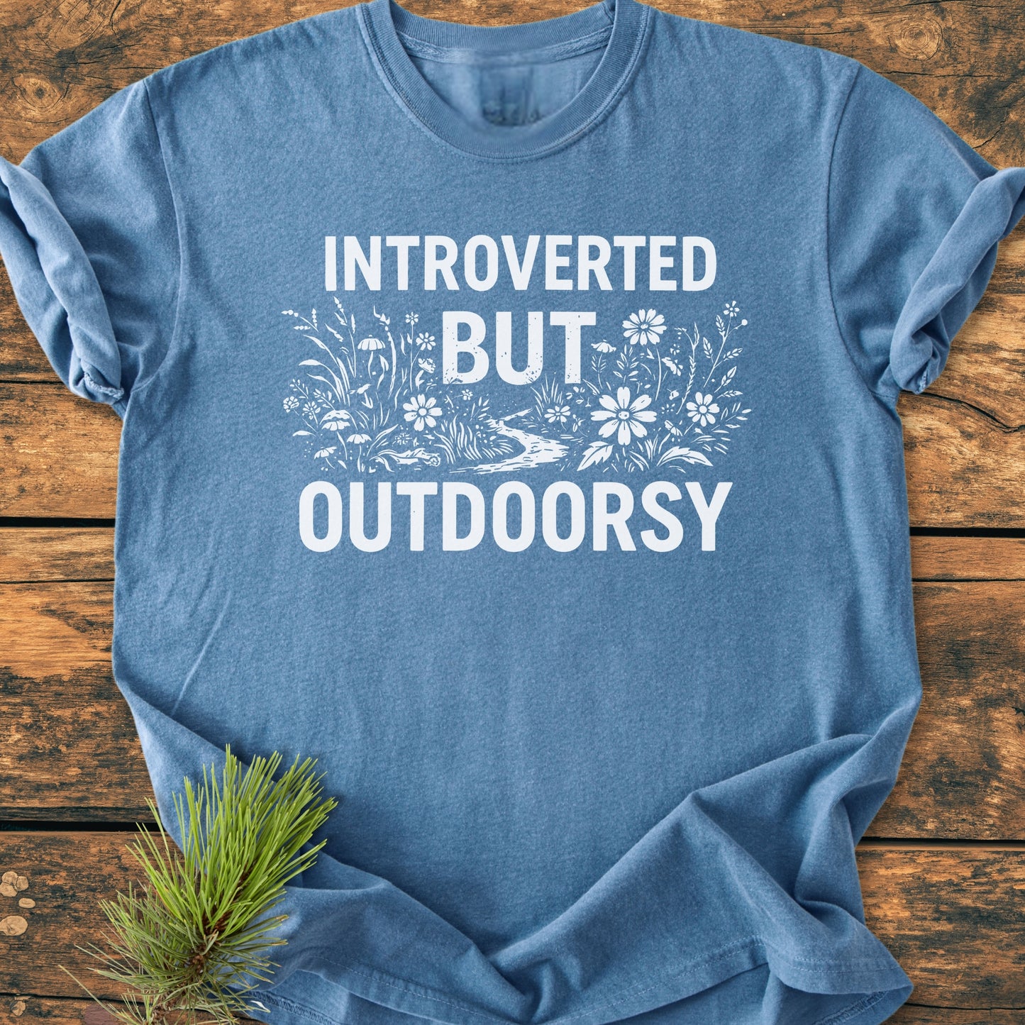 Introverted But Outdoorsy - Vintage Wash Tee features a heather-blue color with a white slogan and nature motif, displayed on wood, accented by a pine sprig, exuding a cozy, relaxed outdoor vibe.