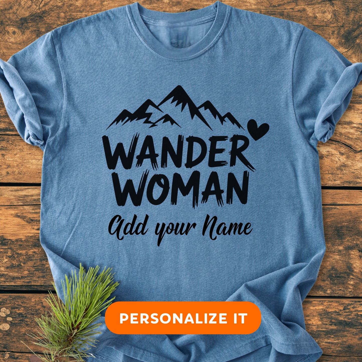 Heather-blue T-shirt with WANDER WOMAN and Add your Name customization option, featuring mountain peaks, a heart, and a “PERSONALIZE IT” button, styled on rustic wood with a pine sprig.