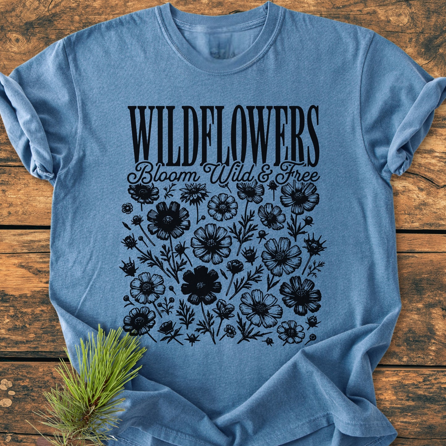 Wildflowers Bloom Wild And Free - Vintage Wash Tee: Light-blue T-shirt with black wildflower graphic and WILDFLOWERS text, laid on wood, featuring a pine sprig nearby. Soft, durable, relaxed fit, 100% ring-spun cotton.