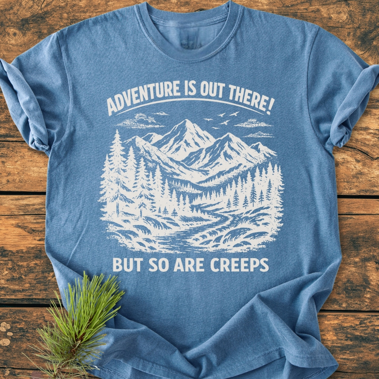 Adventure Is Out There - Vintage Wash Tee in heather-blue, featuring a white mountain and forest scene graphic with playful text, displayed on a wooden surface with a pine sprig beside it.