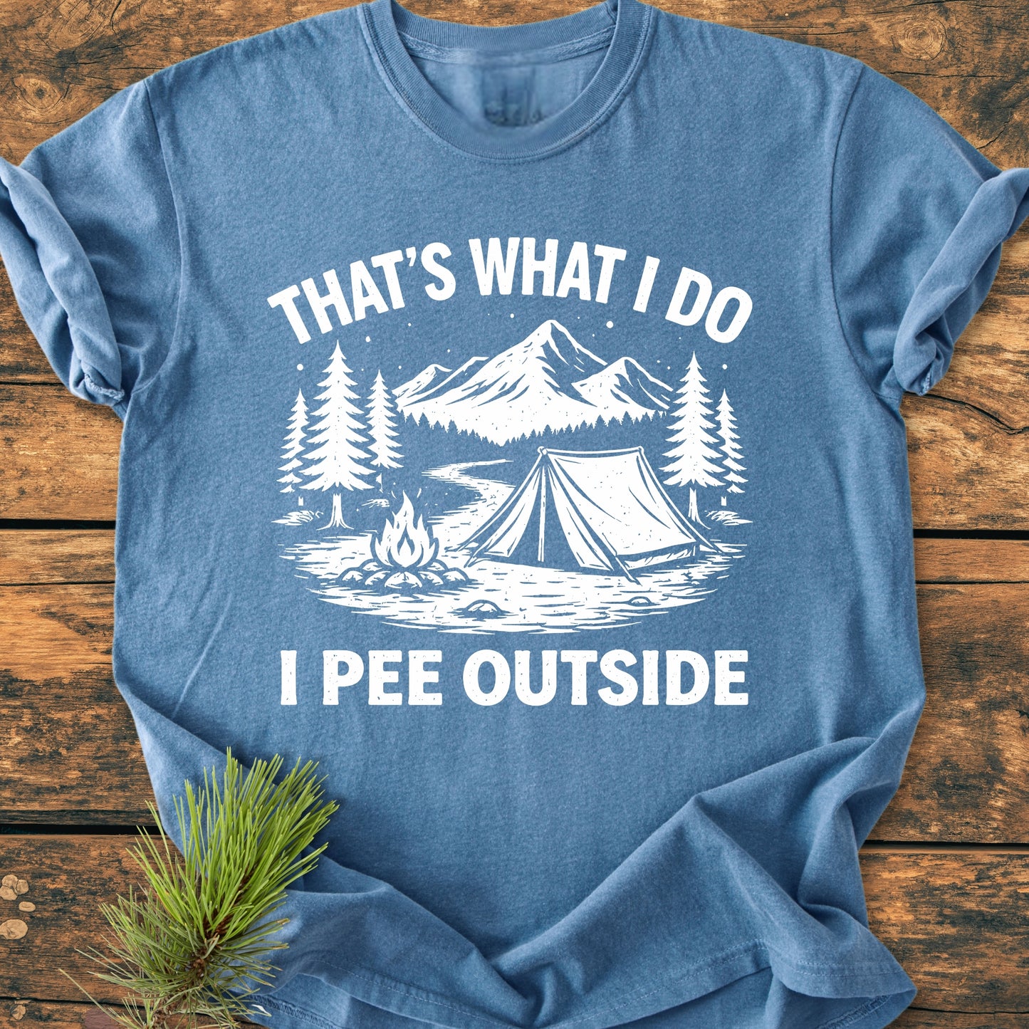 Heather-blue I Pee Outside - Vintage Wash Tee featuring a rustic camp scene with mountains, pine trees, a tent, and a campfire, laid on wood with a pine sprig beside it.