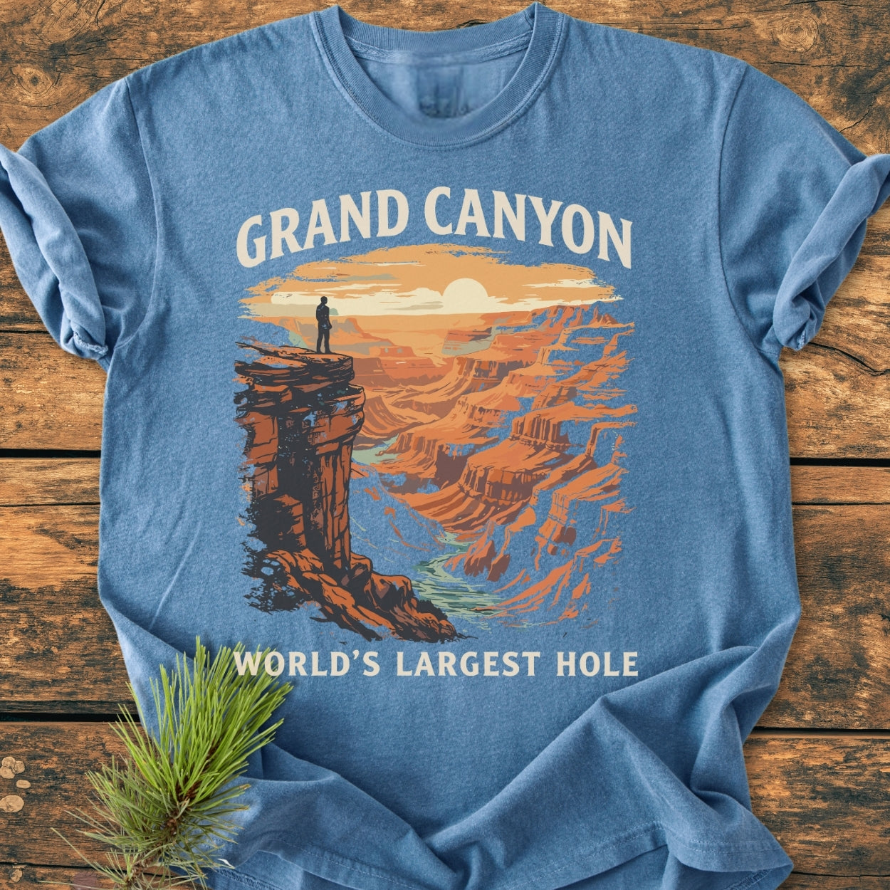 Grand Canyon Large Hole - Vintage Wash Tee featuring a lone figure on a cliff with canyon artwork, styled on wooden surface with pine sprig, highlighting its relaxed fit and soft-washed comfort.