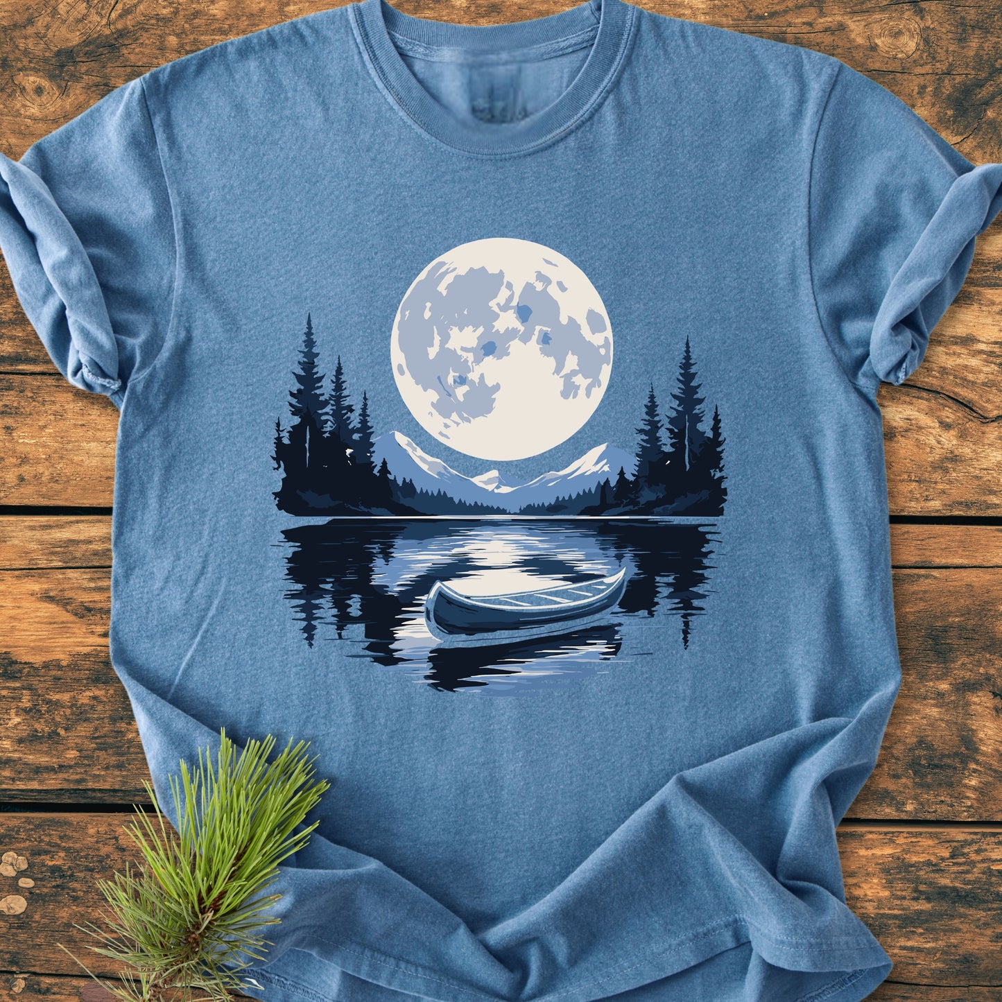 Moonlit Canoe - Vintage Wash Tee features a moonlit wilderness scene with a full moon, mountains, and pine trees reflected in a lake with a canoe, on a soft, relaxed-fit blue t-shirt.