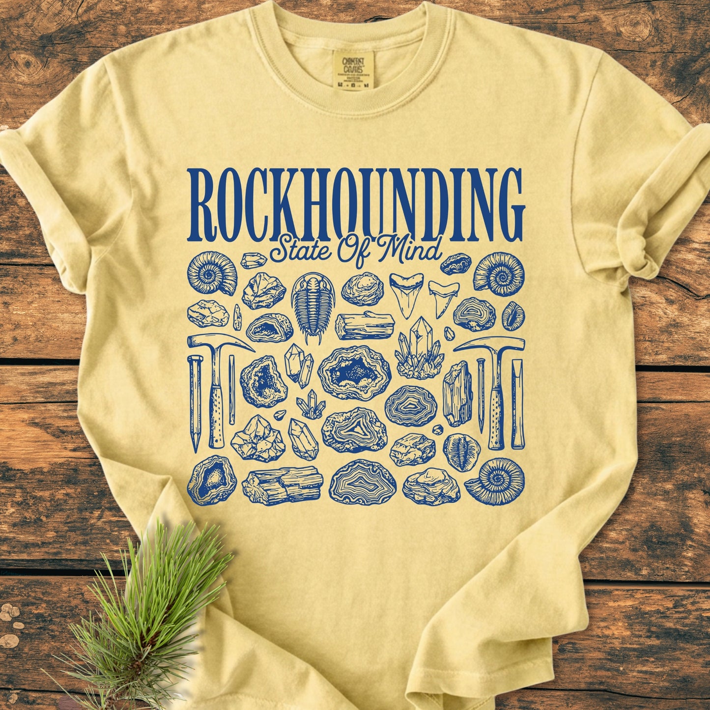 Rockhounding State Of Mind - Vintage Wash Tee featuring hand-drawn rockhounding graphics, laid flat on rustic wood, showcasing its relaxed fit and durable Comfort Colors garment-dyed fabric, with a pine sprig accent.