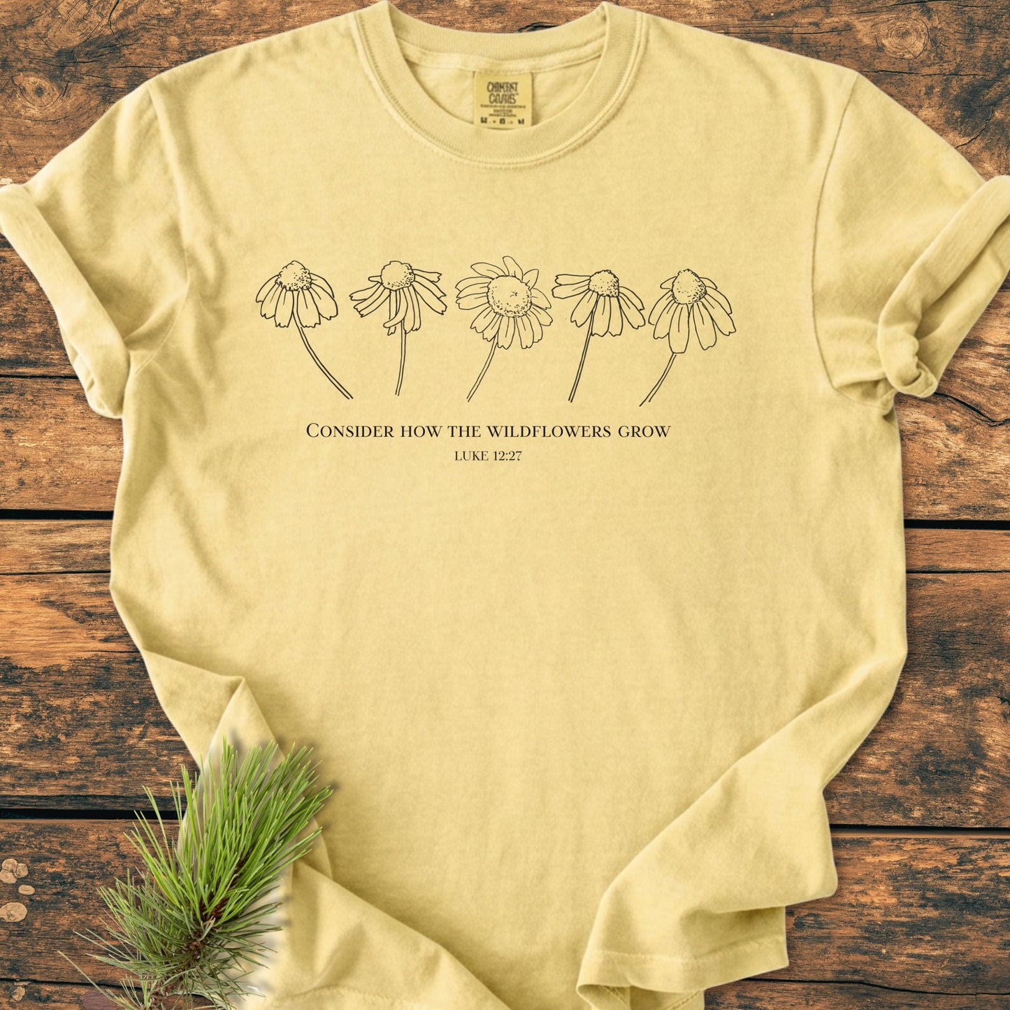 Consider How The Wildflowers Grow - Vintage Wash Tee, featuring a black line drawing of drooping wildflowers and Bible verse on a pale-yellow backdrop, laid on a rustic wooden surface.