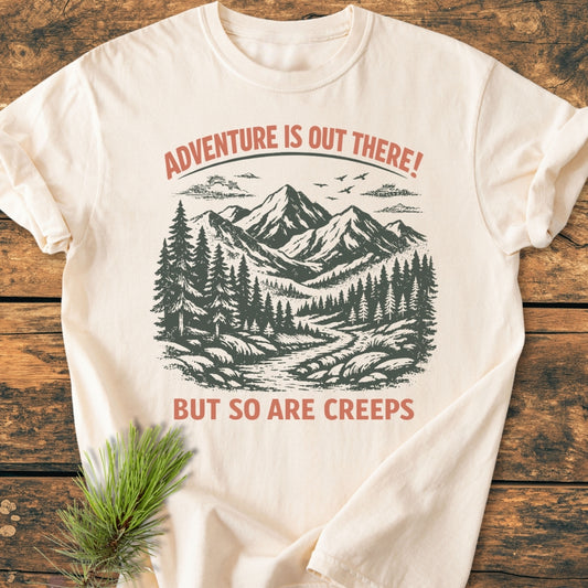 Adventure Is Out There - Vintage Wash Tee featuring a vintage mountain and forest design, laid on wood, with ADVENTURE IS OUT THERE! and BUT SO ARE CREEPS. Pine sprig detail included.