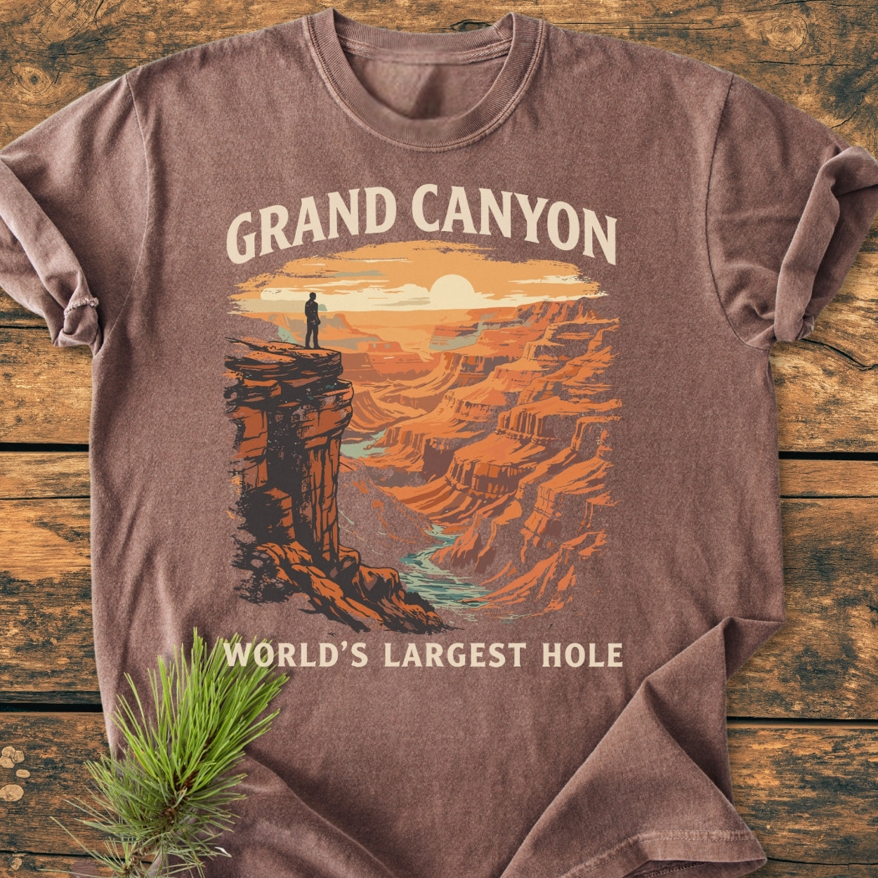 Grand Canyon Large Hole - Vintage Wash Tee features a sunset illustration of the canyon with a silhouette and river. Laid on wood, accompanied by a pine sprig, highlighting its relaxed, durable design.