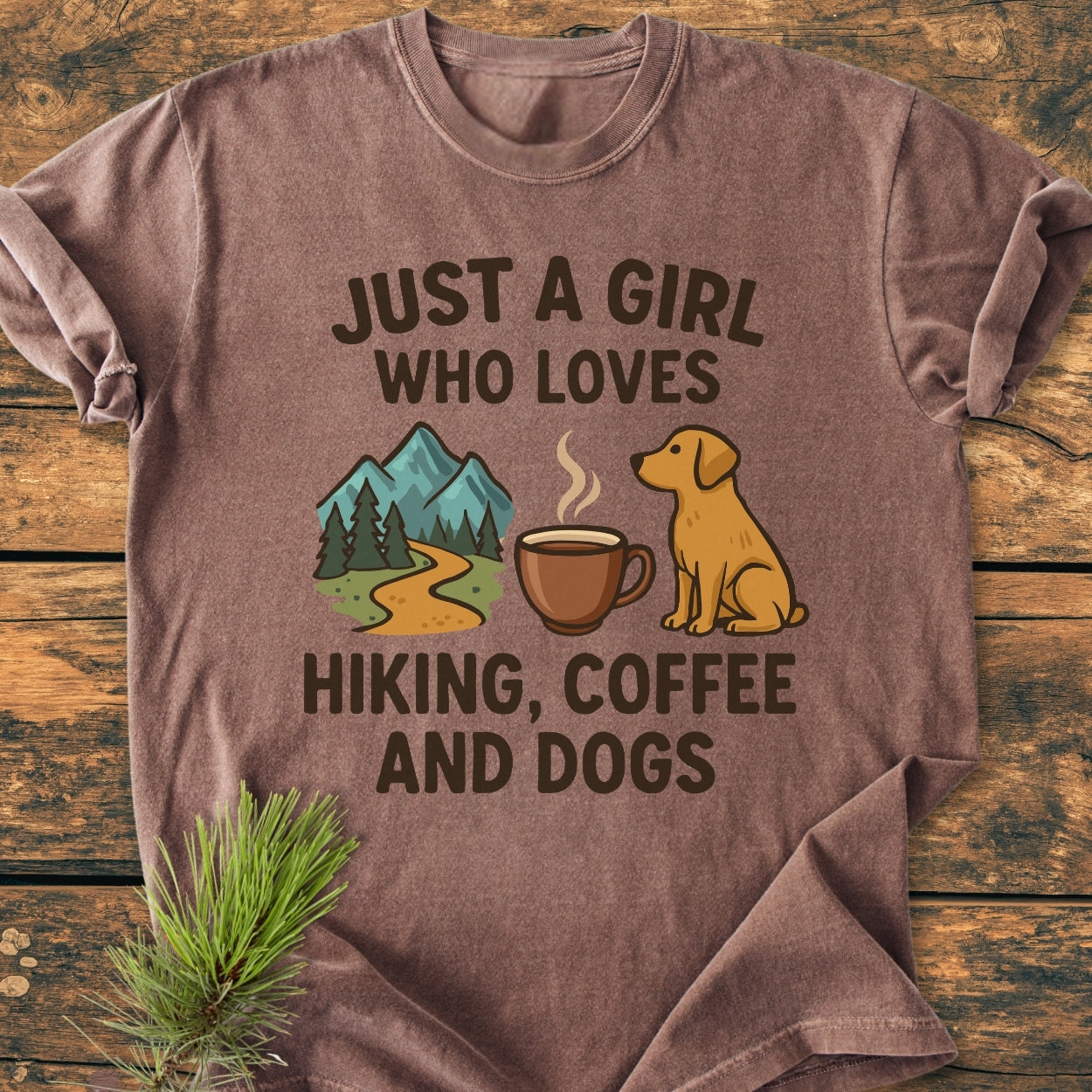 Just A Girl... Hiking Coffee Dogs - Vintage Wash Tee: Brown short-sleeve shirt with hiking, coffee, dog icons on front text. Laid on wood, pine sprig tucked under corner.