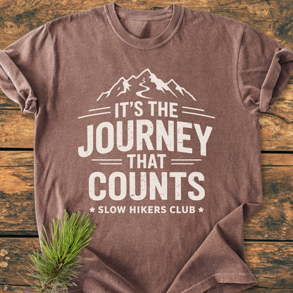 It's The Journey That Counts - Vintage Wash Tee, featuring a white mountain graphic and slogan, folded on a wooden surface with an evergreen sprig, showcasing a relaxed fit and durable stitching.