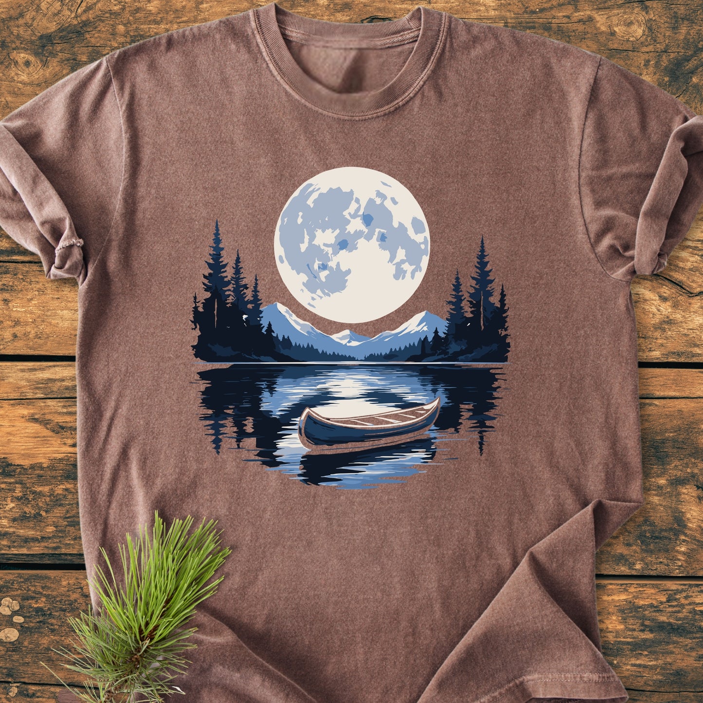 Moonlit Canoe - Vintage Wash Tee: A brown, short-sleeved T-shirt featuring a night wilderness scene with mountains, trees, and canoe, displayed on a wooden surface with a pine sprig.