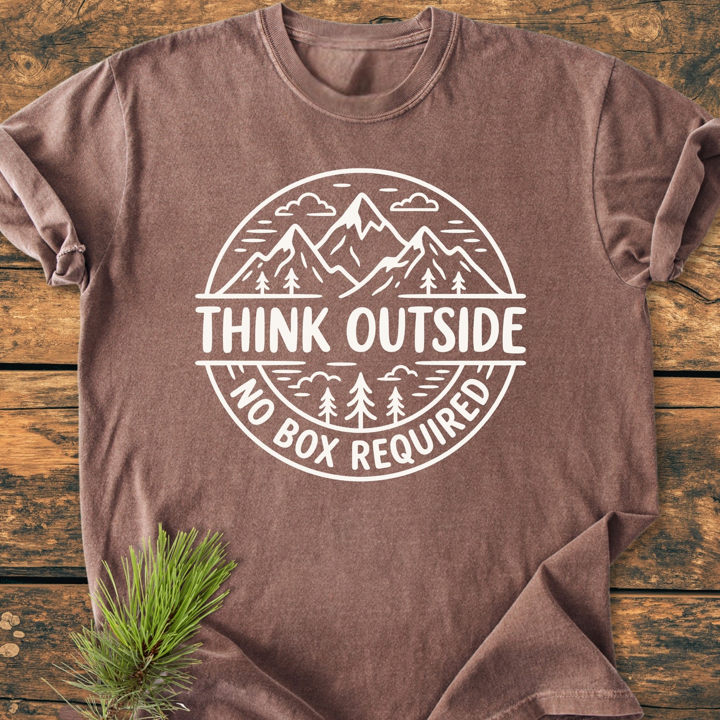 Think Outside - Vintage Wash Tee featuring a white outdoor-themed graphic with mountains and trees, laid flat on a wooden surface with a pine sprig, highlighting its adventure-inspired design.