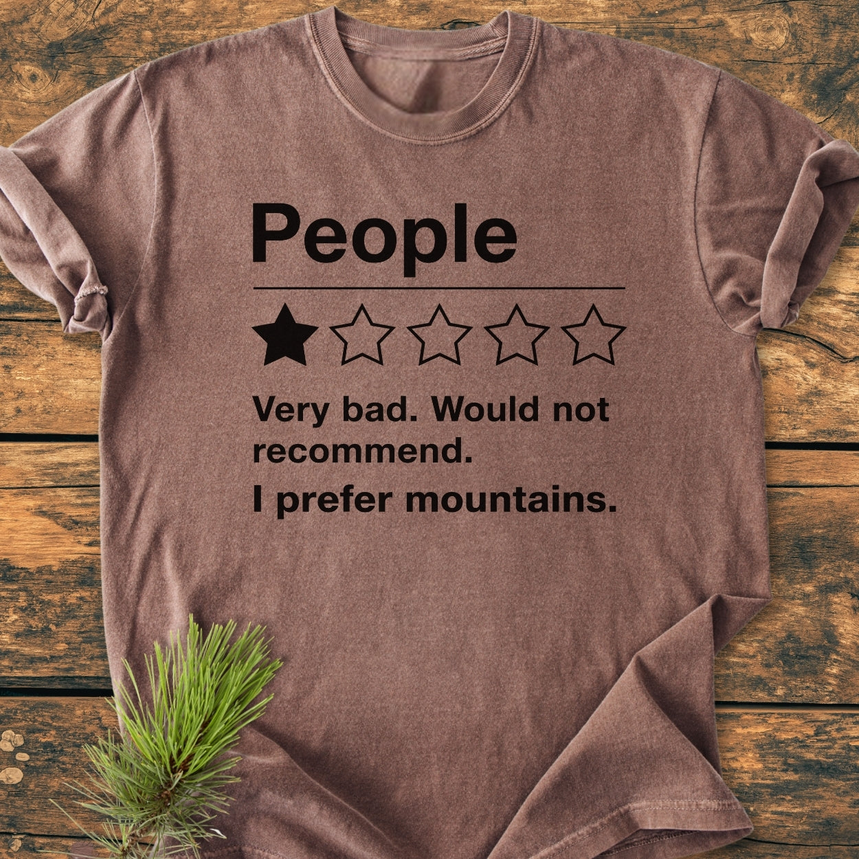 The I Prefer Mountains - Vintage Wash Tee features a playful one-star review design, laid flat on a wooden surface with pine needles tucked under its edge.