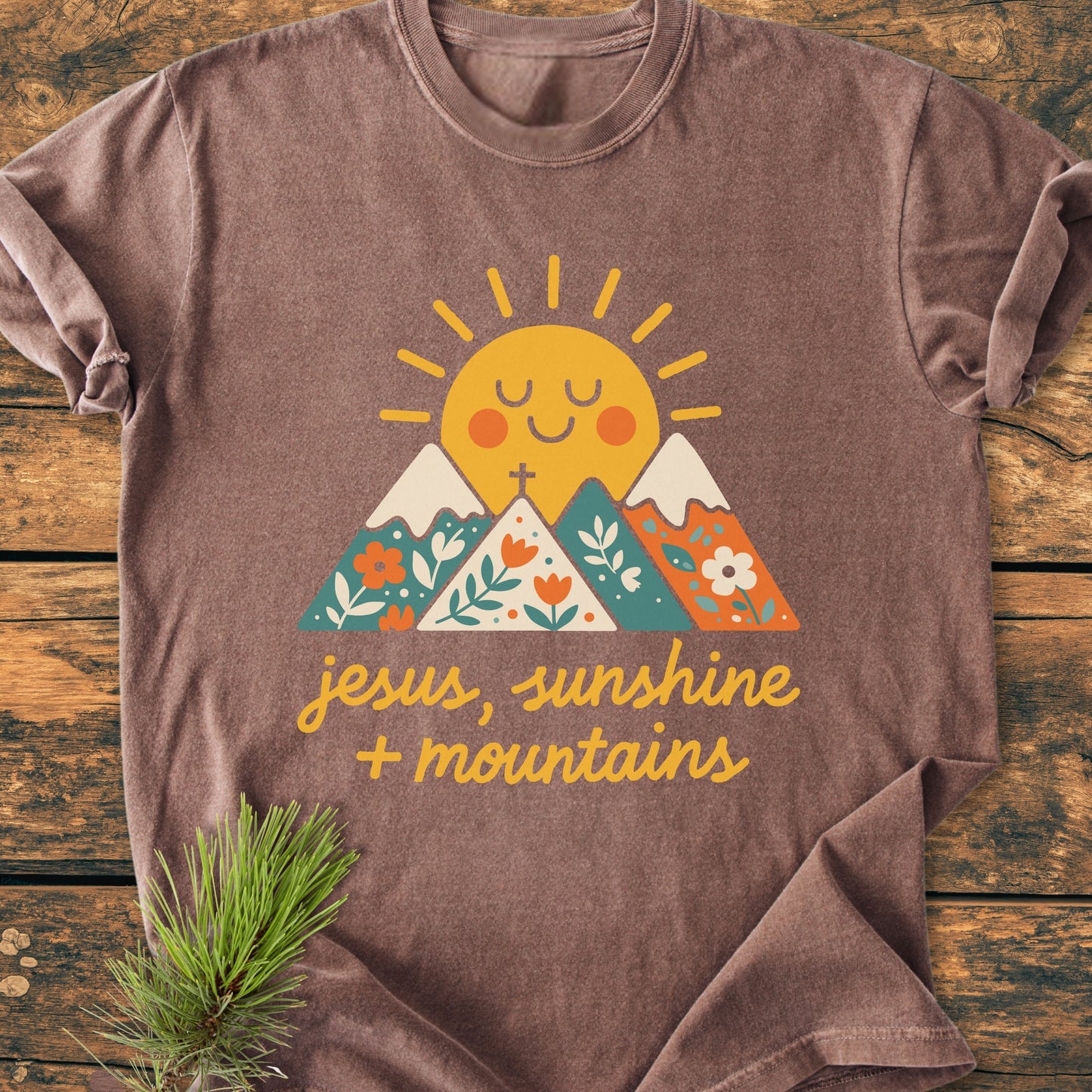 Jesus, Sunshine, Mountains - Vintage Heavyweight Tee: A heather-brown T-shirt featuring a cheerful sun and mountain design with a slogan, laid flat on wood, next to a pine sprig.
