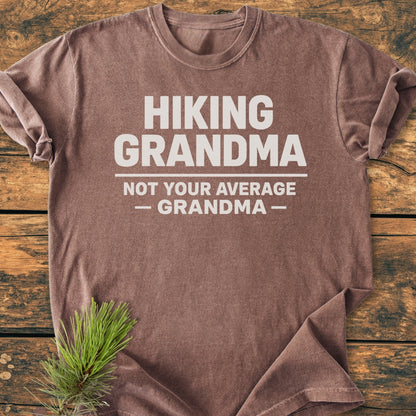 Hiking Grandma - Vintage Wash Tee: Heather-brown T-shirt with bold white slogan, HIKING GRANDMA — Not Your Average Grandma —, displayed on wooden surface, featuring a pine sprig for an outdoorsy touch.