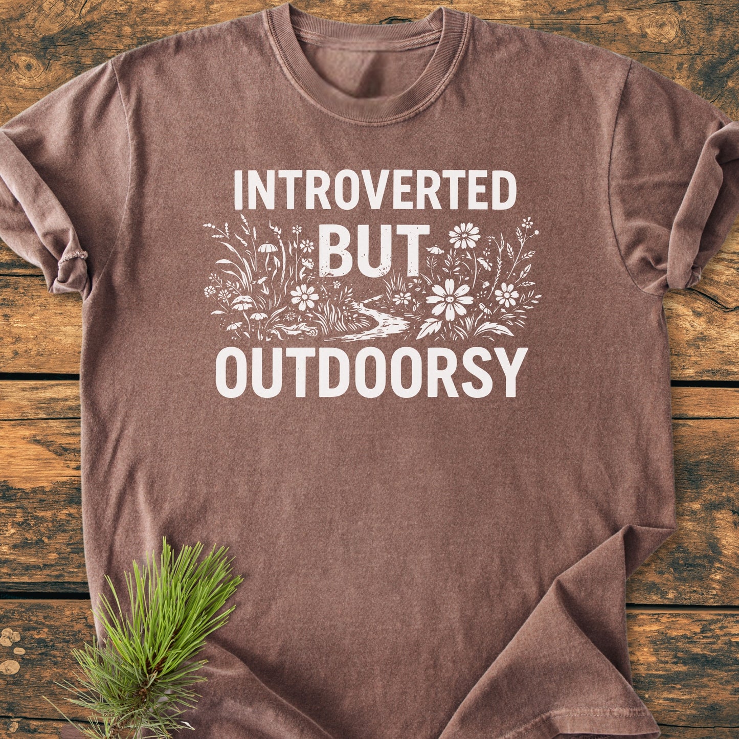 Introverted But Outdoorsy - Vintage Wash Tee features a nature scene and slogan on a brown T-shirt, displayed on wood with a pine sprig, highlighting its rustic, nature-inspired theme.