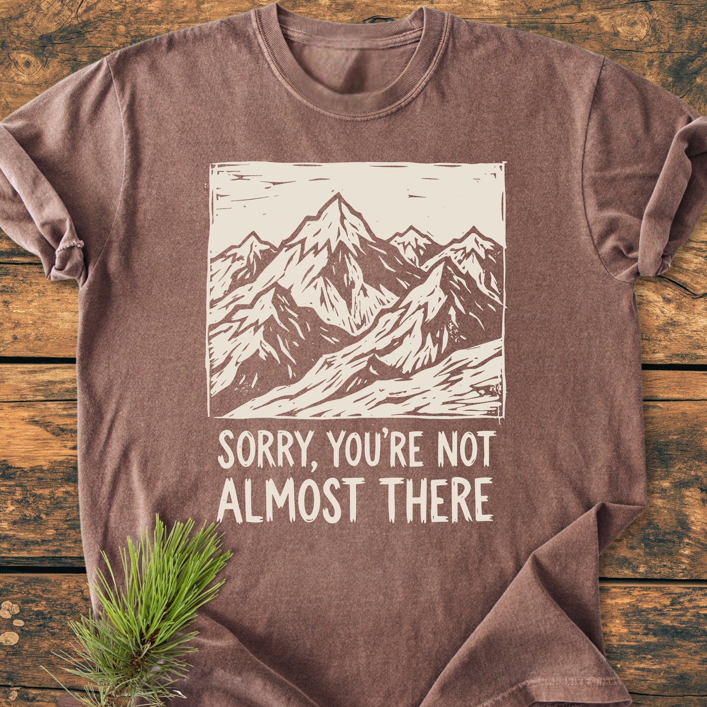 Not Almost There - Vintage Wash Tee features a mountain range line-drawing and slogan on a folded, earth-tone shirt. Laid on wood, it highlights the relaxed fit and durability of the Comfort Colors design.