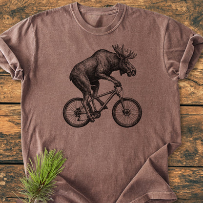 Moose On The Road - Vintage Wash Tee featuring a moose on a bicycle in black line-art on a brown-heather shirt, styled with a pine bough, laid on a rustic wood surface.