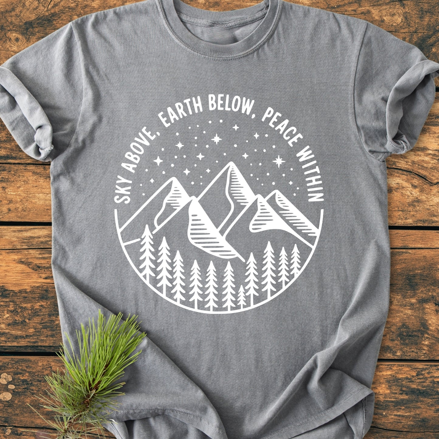 Sky Above - Vintage Wash Tee, featuring a white badge graphic with pine trees, mountains, and stars, displayed on a rustic wooden surface with a pine sprig accentuating its nature-inspired theme.