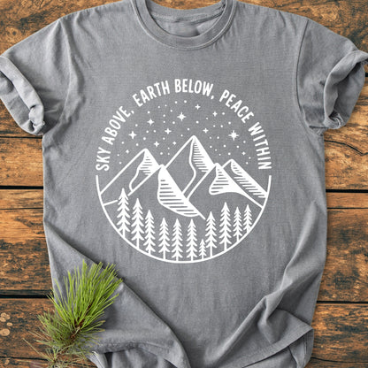 Sky Above - Vintage Wash Tee, featuring a white badge graphic with pine trees, mountains, and stars, displayed on a rustic wooden surface with a pine sprig accentuating its nature-inspired theme.