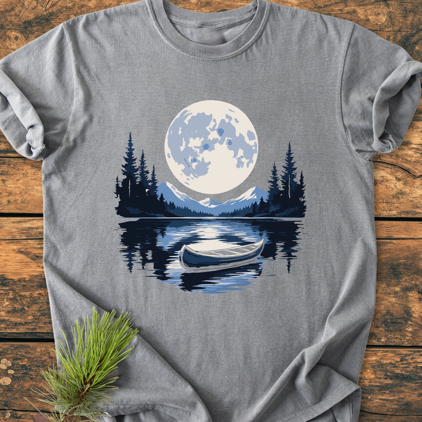 Moonlit Canoe - Vintage Wash Tee features a serene lakeside scene with a full moon, mountains, pines, and a canoe, printed on a heather-gray Comfort Colors shirt laid on wood.