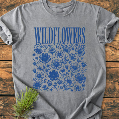 Wildflowers Bloom Wild And Free - Vintage Wash Tee featuring bold blue WILDFLOWERS text and wildflower silhouettes on a heather-gray background, laid on rustic wood with pine needle accent.