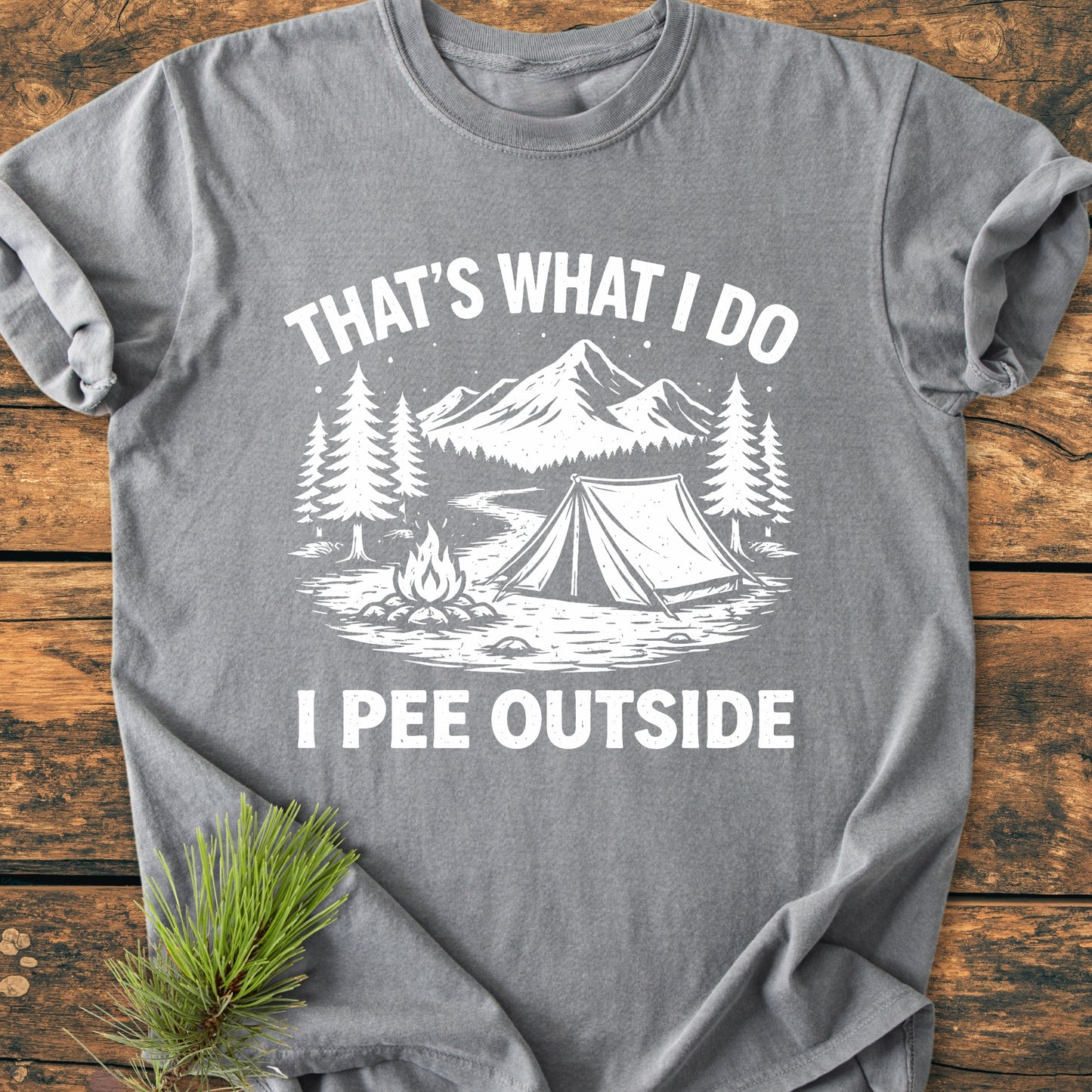Heather-gray I Pee Outside - Vintage Wash Tee featuring a stylized camping scene with pine trees, mountains, a tent, and campfire, laid flat on a wooden surface, highlighting its relaxed, soft-washed design.