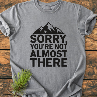 Sorry You're Not Almost There - Vintage Wash Tee in grey with a bold mountain silhouette and text. Laid flat on wood, with a pine sprig accentuating its relaxed, durable design.