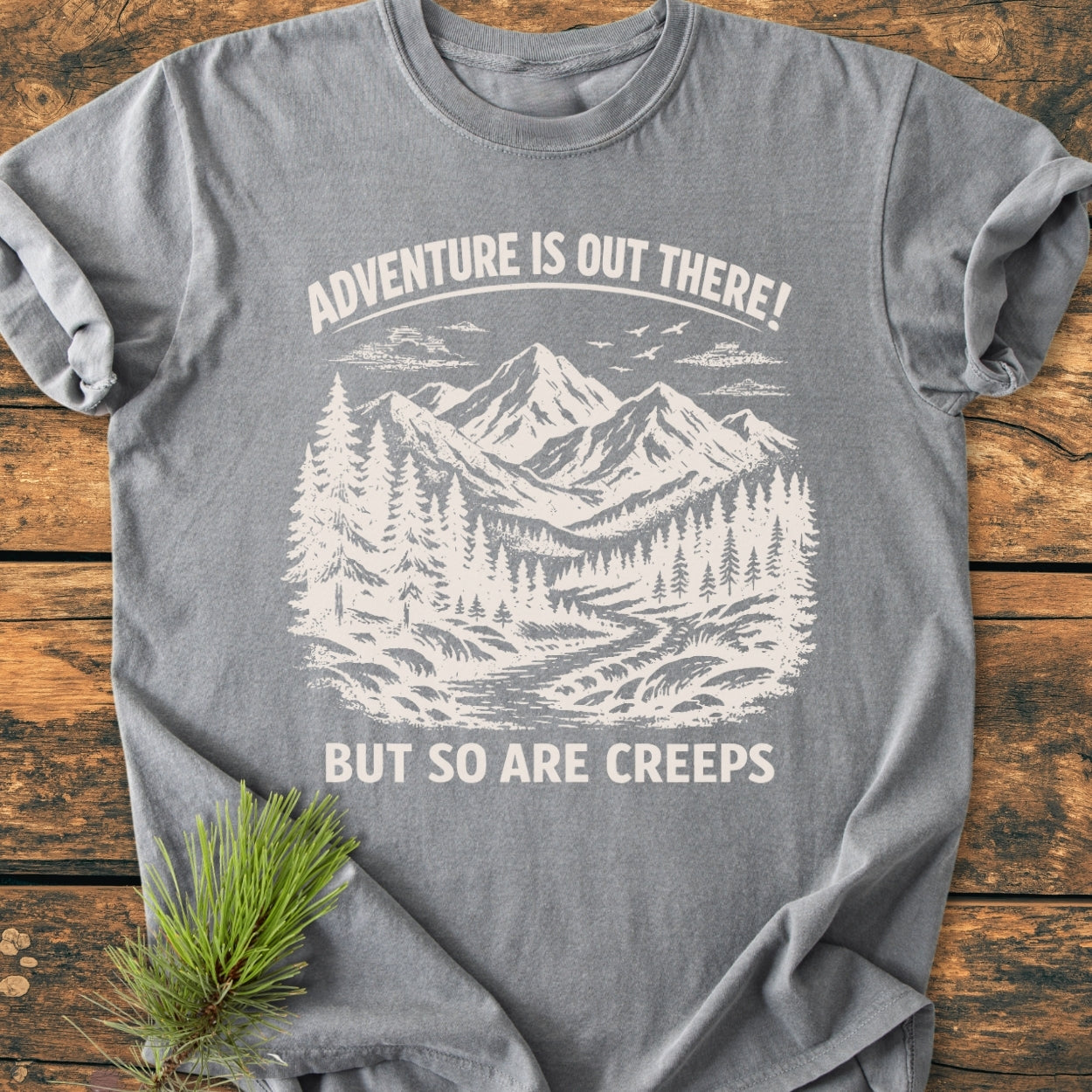 Adventure Is Out There - Vintage Wash Tee featuring a white woodcut-style illustration of mountains, pine trees, and a stream, laid flat with a pine sprig accent on a wooden surface.
